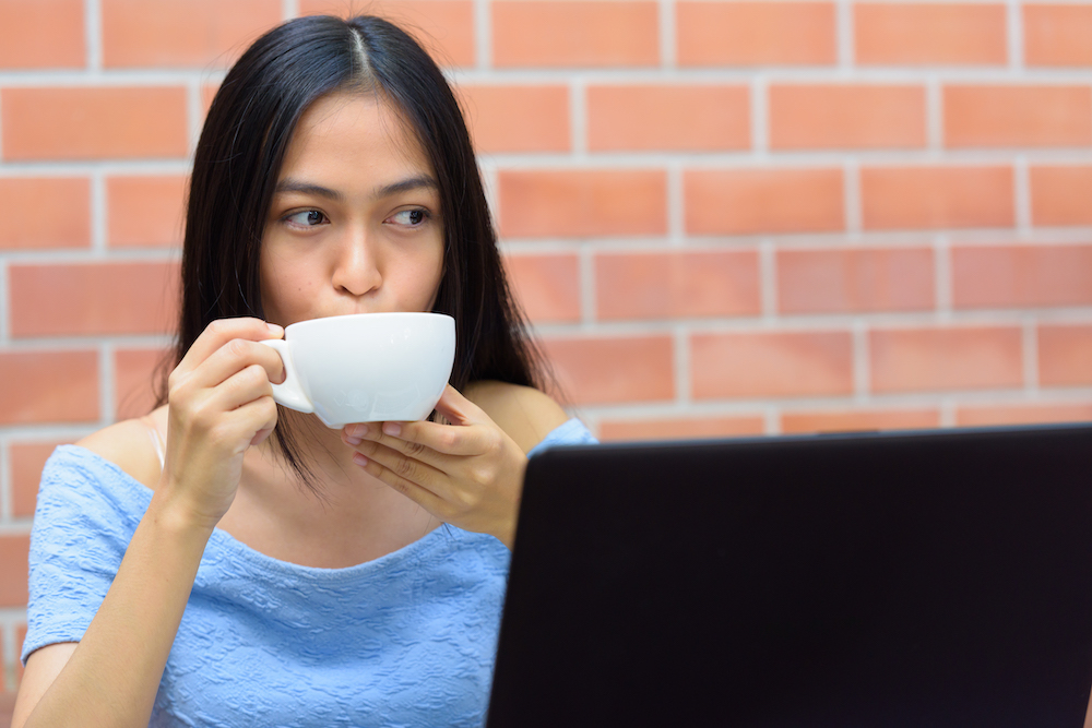 Una adolescente asiática bebiendo café mientras piensa con una laptop contra una pared de ladrillos