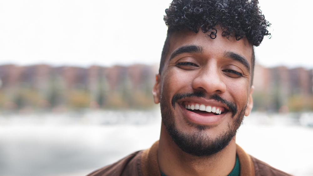 Retrato de hombre latino feliz sonriendo y mirando a la cámara al aire libre con la ciudad en segundo plano.